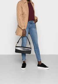 Gequiltede beige jas, bordeaux top, blauwe jeans, zwarte clutch met gouden ketting, en zwarte sneakers met klittenbandsluitingen.