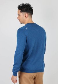 Pull en tricot bleu à manches longues, col rond et ourlet côtelé. Présente une petite écusson avec logo sur l'épaule arrière.