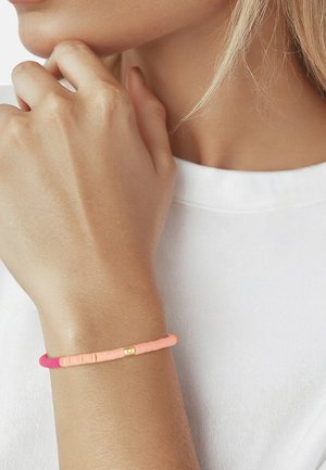 Handgelenk mit einem schlanken Perlenarmband in Rosa- und Koralltönen bei einer Person, die ein weißes Hemd trägt, Hand nahe am Gesicht.