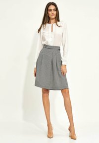 Kobieta w białej bluzce z długim rękawem i czarno-białej spódnicy w zygzaki do kolan z wysokim stanem, w przezroczystych szpilkach.