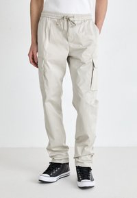 Beige Cargo-Hosen verfügen über einen elastischen Bund mit Kordelzug, seitliche Taschen und schmal zulaufende Beine, kombiniert mit schwarzen High-Top-Sneakern.