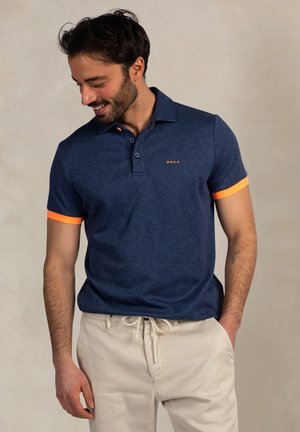 Man die glimlacht en naar beneden kijkt, draagt een marineblauw poloshirt met oranje mouwboorden en beige broek met trekkoord, staand voor een effen achtergrond.