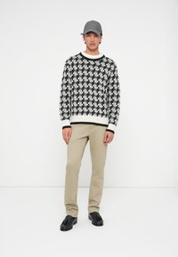 Čierno-biely pletený sveter s geometrickým vzorom, béžové nohavice a čierne mokasíny. Šedá čiapka dokončuje outfit.