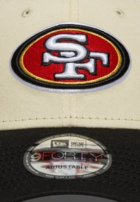 Șapcă de baseball bej, cu un logo brodat roșu și negru, cu "SF" în centrul său. Vârf negru, curea ajustabilă și branding New Era pe partea inferioară.