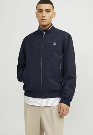 Chaqueta bomber azul marino con cuello alto, cierre de cremallera y bolsillos laterales. Presenta un pequeño parche del logo y detalles de rayas en contraste.