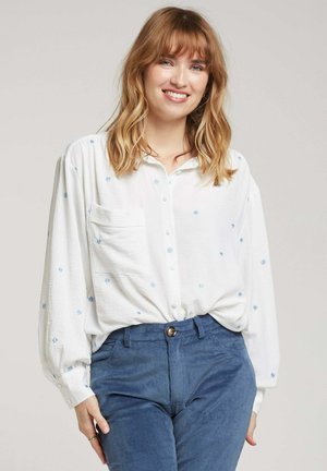 Chemise blanche à boutons avec broderie florale bleue, poche poitrine unique et manches ballon, associée à un pantalon en velours côtelé bleu.