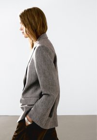 Donna in un blazer grigio e pantaloni scuri con le mani in tasca, rivolta a sinistra su uno sfondo bianco semplice.