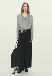 Vrouw met golvend haar die een grijze, gedrapeerde trui draagt, brede zwarte broek, zwarte riem, zwarte laarzen en een grote zwarte tas met structuur vasthoudt.