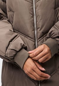 Storos pūkinės striukės ruda spalva su siūtais raštais, sidabrinė priekinė užtrauktukas, ribotos rankogaliai ir laisvas modelis.