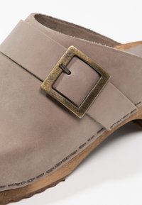 Sabots en suède gris avec une boucle en bronze carrée, des coutures détaillées et une semelle en bois naturel. Le design arrondi met en avant le confort et la simplicité.