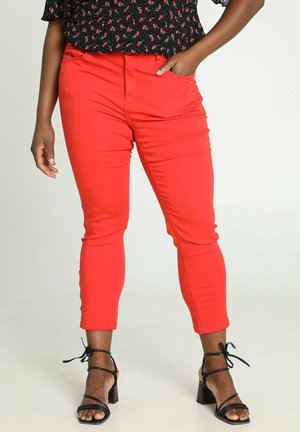 Person wearing slim red pants and black strappy block heel sandals, hand in pocket, standing on light background.