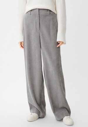 Person, der bærer vidt ben grå kordfløjl bukser, hvid sweater med lange ærmer og hvide sneakers mod en ensfarvet baggrund.