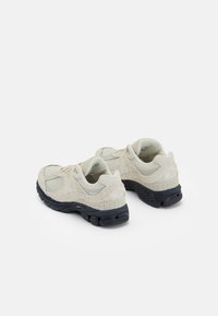 Beige sportcipők sima és texturált felületek kombinációjával, fekete gumitalppal és diszkrét logóval a saroknál.