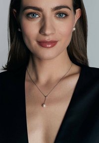 Srebrny naszyjnik z pojedynczym wiszącym perłą, noszony przez modelkę w czarnej bluzce z dekoltem w serek. Gładka tekstura, neutralne tony i minimalistyczny design.