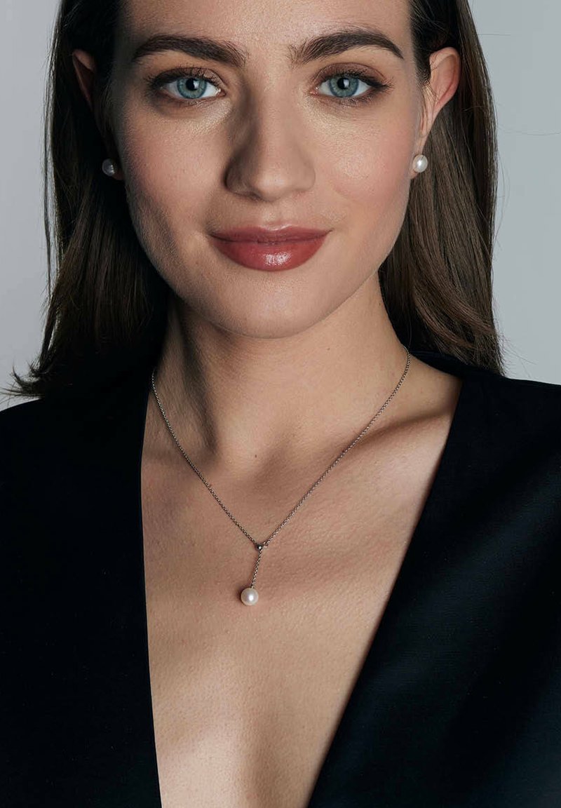Srebrny naszyjnik z pojedynczym wiszącym perłą, noszony przez modelkę w czarnej bluzce z dekoltem w serek. Gładka tekstura, neutralne tony i minimalistyczny design.
