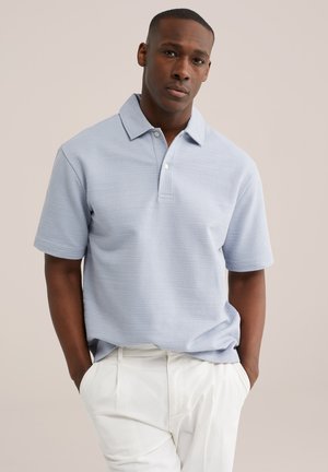 Muž stojaci s rukami v kapse, oblečený v svetlomodrej polokošeli s krátkym rukávom a bielych nohaviciach na jednoduchom pozadí.