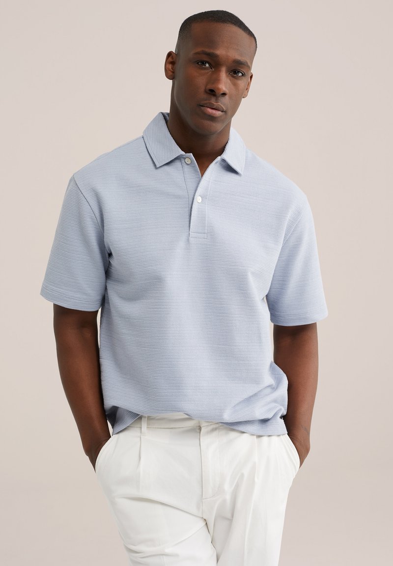 Uomo in piedi con le mani in tasca che indossa una polo a maniche corte azzurro chiaro e pantaloni bianchi su sfondo semplice.