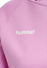 Primo piano di una felpa rosa con logo bianco "hummel" stampato sul petto, che mostra la trama del tessuto e i dettagli delle cuciture.