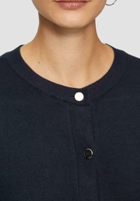 Marineblauer Cardigan mit ribbeltem Kragen, Knopfleiste vorne mit metallischem Akzent und gewebter Textur. Nahaufnahme über der Brust.