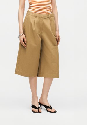 Nő, aki teve színű, bőszárú, térdig érő rövidnadrágot, csíkos narancs-fehér felsőt, fekete magas sarkú szandált és ezüst karkötőt visel.