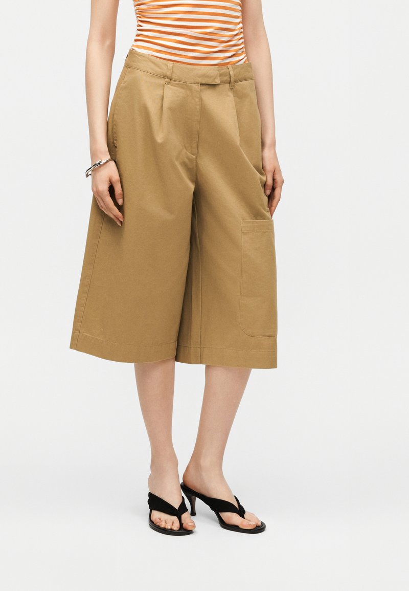 Γυναίκα που φοράει καμηλό φαρδιά σορτς μέχρι το γόνατο, ριγωτή πορτοκαλί και λευκή μπλούζα, μαύρα πέδιλα με τακούνι και ασημένιο βραχιόλι.