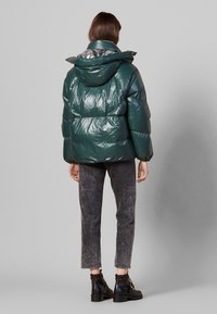 Giacca puffer verde lucido con cappuccio, con foderato a motivo, abbinata a pantaloni grigi e stivaletti neri.