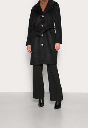 Kobieta ubrana w czarny płaszcz z paskiem i czterema guzikami, czarne spodnie o szerokich nogawkach, czarne spiczaste botki oraz brązowy golf.