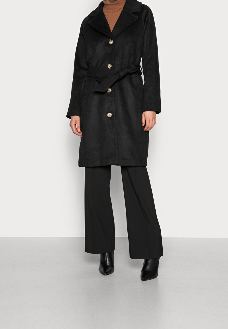 Kvinde iført en sort bæltefrakke med fire knapper, sorte vidde bukser, sorte spidse støvler og en brun højhalset sweater.