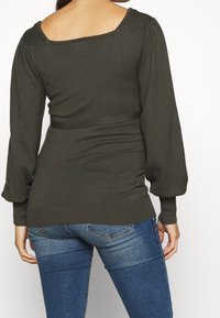 Maglietta verde oliva a maniche lunghe con un ampio scollo a barchetta, spalle a sbuffo e finitura texturizzata; abbinata a jeans in denim blu.