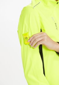 Leuchtend gelbe Jacke mit reflektierendem Design, ausgestattet mit einer schützenden Tasche für einen gelben Power-Knopf am linken Ärmel.