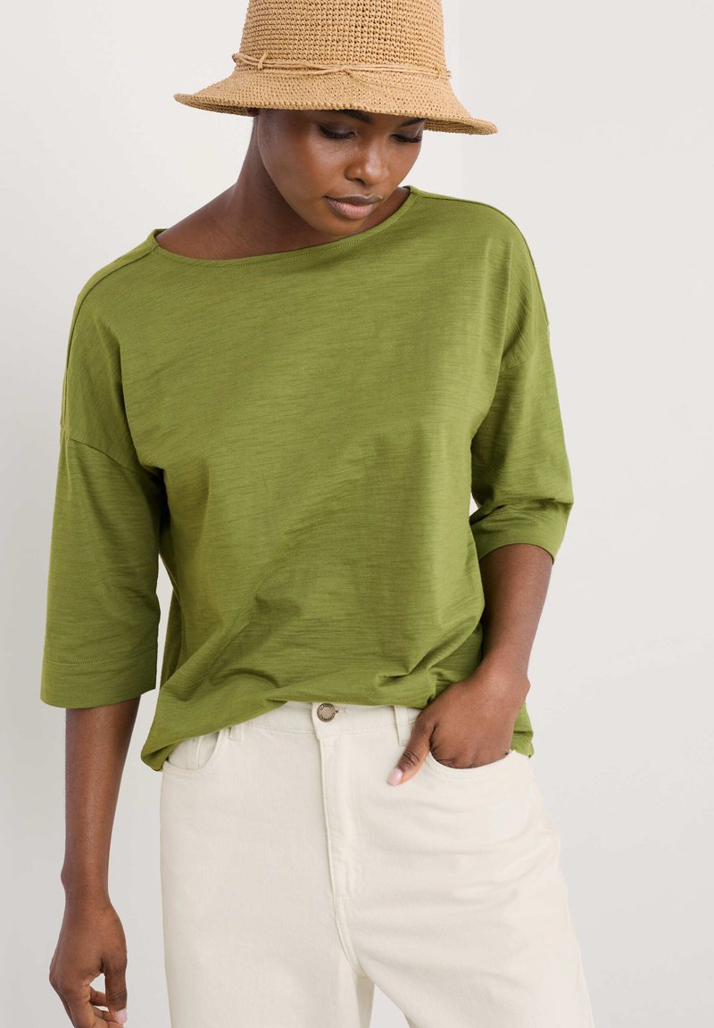 Olive green, loose-fitting top with three-quarter sleeves, paired with light cream pants. Soft texture, minimalist design, no patterns or accents.