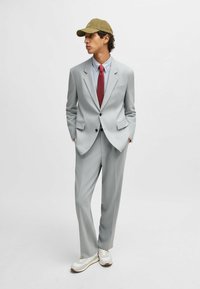 Light gray suit with a fitted blazer and straight-leg trousers. Paired with a light blue shirt, red tie, and an olive green cap. White sneakers.