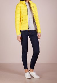 Giacca a vento gialla con cappuccio, presenta un design trapuntato; maglione grigio sottostante, abbinato a jeans skinny scuri e sneakers bianche.