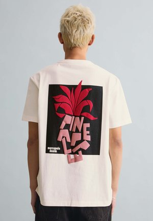 Personne aux cheveux blonds courts portant un t-shirt blanc avec un motif d'ananas rouge et un texte rose au dos, sur un fond neutre.