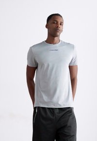 Tricou scurt cu mânecă, de culoare gri deschis, realizat dintr-un material neted, având un mic logo al brandului Calvin Klein în nuanță de albastru pe piept și guler rotund.