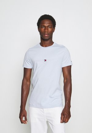 Man die een lichtblauw Tommy Hilfiger T-shirt en een witte broek draagt, staand tegen een effen witte achtergrond.