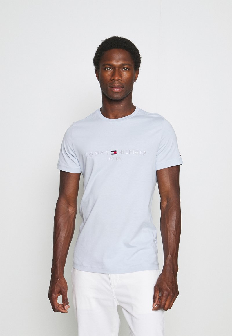 Hombre con camiseta azul claro de Tommy Hilfiger y pantalones blancos posando sobre un fondo blanco liso.
