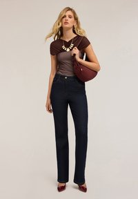 Braun und graues Kurzarmoberteil, das über dunklen Jeans lagiert ist. Das Model trägt eine bordeauxrote Handtasche und trägt dunkelrote spitz zulaufende Schuhe.