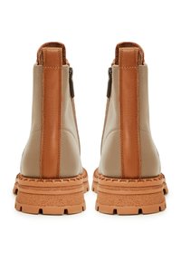 Bottes chelsea en cuir beige avec des accents marron, vue de dos. Présente une semelle texturée et des zips sur le côté. Design robuste et imposant.