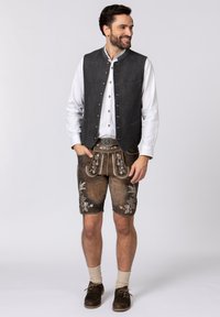 Graue Weste mit metallischen Knöpfen über einem weißen Hemd, kombiniert mit braunen bestickten Ledershorts und hellen Socken.