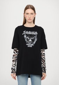 Svart oversized t-skjorte med trykk av en panter og teksten "Sauvage Versace Jeans Couture." Lange ermer med zebra-mønster under.