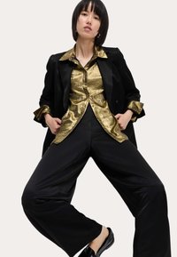 Blazer negro sobre una camisa dorada brillante, combinado con pantalones negros holgados y mocasines negros, sobre un fondo neutro.