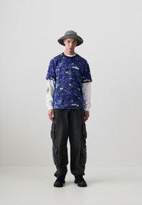 Purple-patterned short-sleeve shirt over a white long-sleeve top, paired with dark cargo pants. Accessorized with a grey hat and black shoes.