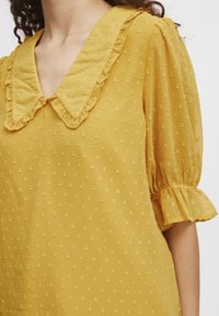 Gelbe Bluse mit Rüschenkragen, kurzen Puffärmeln und einem strukturierten Punktmuster im gesamten Stoff. Leichtgewichtiges und durchscheinendes Material.