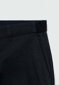 Pantalon noir avec poche latérale et détail passant de ceinture sur un fond clair uni, montrant la partie haute de la taille.