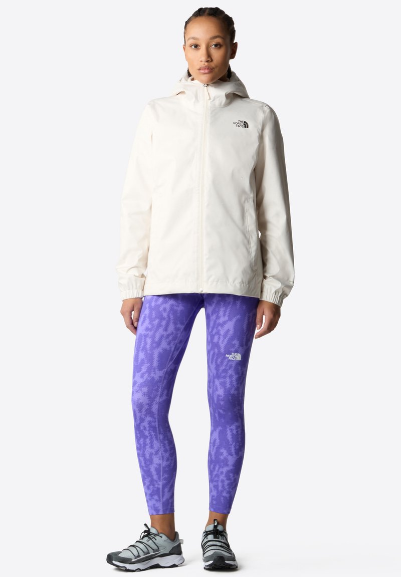Valkoinen vedenpitävä takki huppuineen, yhdistettynä purppuran värisiin kuviollisiin legginseihin. Asusteena harmaat urheilukengät.