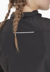 Sort jakke med en tekstureret overflade, der har en høj krave og en reflekterende hvid detalje nær ryggen af halsudskæringen.