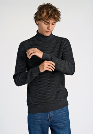 Dunkelgrauer, gestrickter Pullover mit hohem Kragen, der ein strukturiertes Muster und gerippte Bündchen aufweist. Kombiniert mit blauen Jeans.