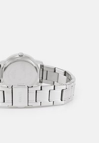 Stainless steel watch with a round case, silver metal bracelet, and brushed finish. Clasp features "GUESS" logo. Visible movement on the back.