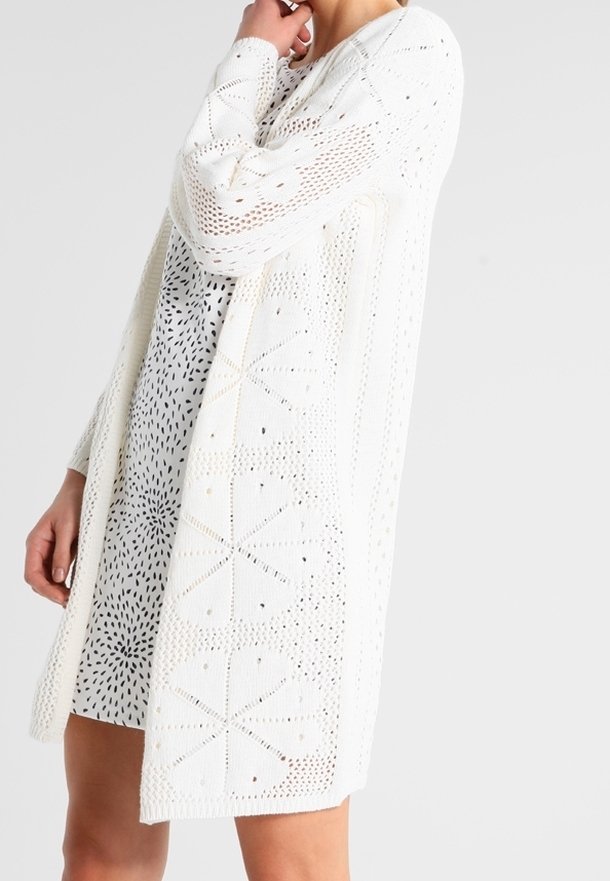 Femme portant un cardigan blanc en maille ouverte sur une robe blanche à motif abstrait en points noirs, debout devant un fond clair uni.